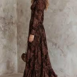 May include: A long, brown floral print dress with long sleeves and a ruffled hem. The dress has a collared neckline and is paired with brown sandals and a small, round purse.