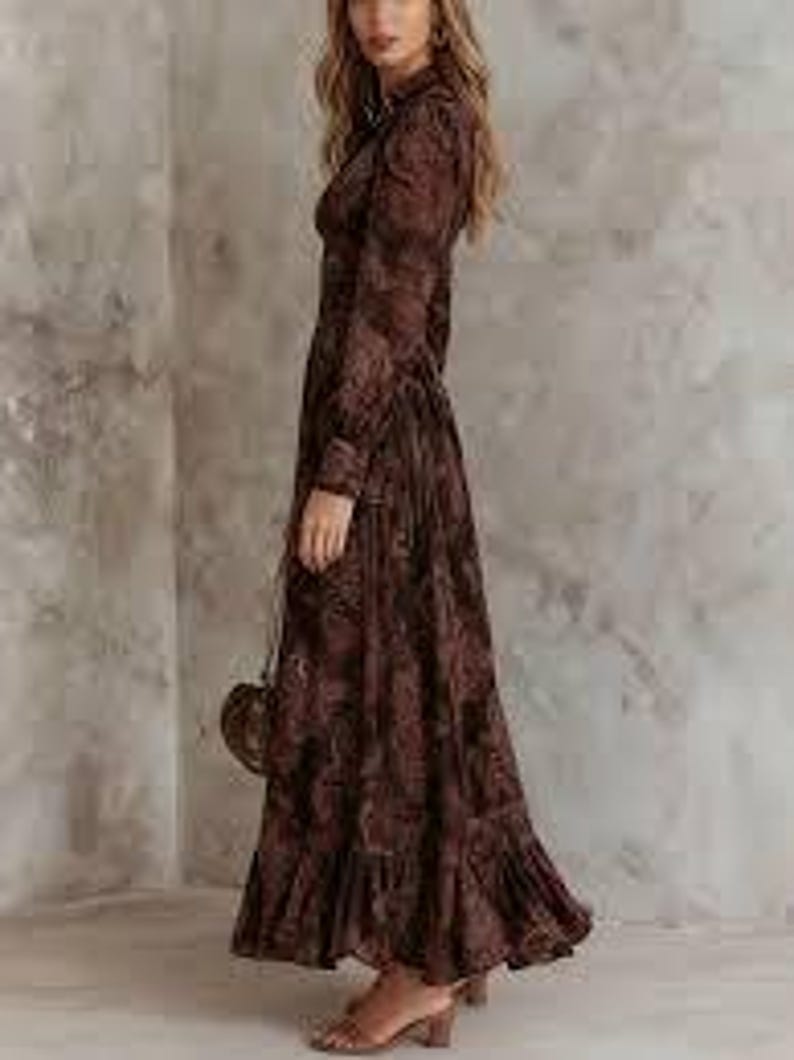 May include: A long, brown floral print dress with long sleeves and a ruffled hem. The dress has a collared neckline and is paired with brown sandals and a small, round purse.