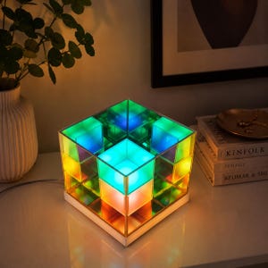 Peut inclure: Une lampe de table carrée composée d'un cube en acrylique translucide multicolore. La lampe émet une lueur vive dans des tons de vert, bleu et orange, créant une illusion d'optique de profondeur grâce à sa structure réfléchissante en grille.