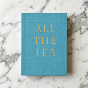 May include: A teal-colored book with the words "ALL THE TEA" in gold lettering. The book is on a white and gray marble surface. The book has a textured cover.
