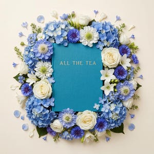 May include: A teal book with gold lettering reading "ALL THE TEA" is surrounded by a wreath of blue and white flowers. The wreath includes hydrangeas, roses, and other floral elements, creating a vibrant, decorative arrangement.