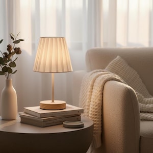 May include: A small, beige pleated lampshade sits atop a light wood base and stem. The lamp is on a round, gray table with books and a vase of greenery. A cream-colored armchair and blanket are in the background.