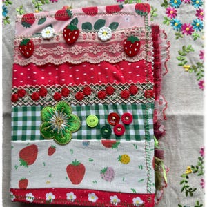 Op de afbeelding: Een handgemaakt stoffen tijdschrift met een aardbeienthema. De omslag heeft lagen van roze, rode en witte stoffen, versierd met kant, knopen en geborduurde details. Het tijdschrift is versierd met aardbeien- en bloempatronen.