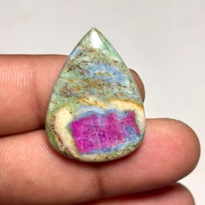 Peut inclure: Une pierre précieuse polie en forme de goutte d'eau présentant un centre magenta vibrant entouré de bandes bleues, crème et vert clair. La pierre a une surface lisse et brillante avec des inclusions minérales naturelles et un motif distinct.