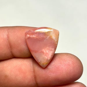 May include: A triangular-shaped pink gemstone with a smooth, polished surface. The stone features translucent and opaque sections with subtle white and pale yellow marbling throughout its structure, showcasing a natural, glossy finish.