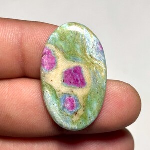 May include: An oval-shaped cabochon gemstone featuring a polished surface with a mix of vibrant magenta-pink inclusions set against a variegated background of pale cream, light green, and soft blue-grey mineral patterns throughout the stone.
