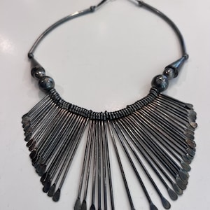 May include: A dark silver-toned metal collar necklace featuring a rigid circular neck wire that transitions into a wide, fan-shaped fringe of thin, rod-like pendants. Each rod ends in a small, flattened, rounded tip, with two larger spherical beads.