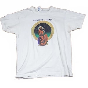May include: White t-shirt featuring the text "Grateful Dead" above a colorful graphic. The graphic depicts a robed figure within a circular frame against a starry background. The shirt has short sleeves and a crew neck.