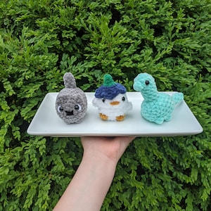 May include: Three small crocheted plush toys on a white rectangular tray. The left toy is a round gray creature with a small tail. The center toy is a white bird with a blue hat. The right toy is a light green long-necked dinosaur figure.