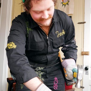 May include: A person wearing a green crochet hat with white shamrocks, black jumpsuit with embroidered designs, and work gloves, using a hammer on a wooden plank. The jumpsuit has the name "Jack" embroidered on it.