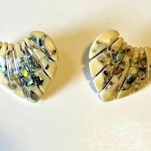 May include: A pair of heart-shaped earrings. Each earring is cream-coloured with inlaid iridescent pieces. The earrings are segmented, with each segment following the heart's curve. The iridescent pieces reflect various colours.