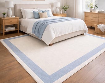 May include: A bedroom scene featuring a large bed with white bedding and a light blue throw blanket. A cream-colored rug with a light blue border is on the wooden floor. The room has a neutral color palette with natural light.