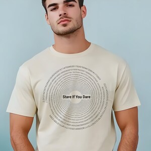 May include: A cream-colored t-shirt featuring a circular design of numbers and the phrase "Stare If You Dare" in the center. The shirt is a classic crew neck style.