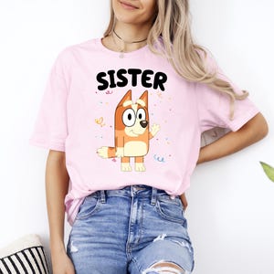 May include: A light pink t-shirt with the word "SISTER" in black block letters. The shirt features a cartoon dog character in shades of orange, tan, and white. The dog is waving, and there are colorful confetti accents.