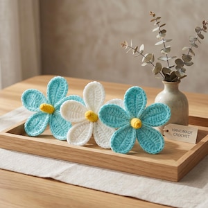 Puede incluir: Tres flores de ganchillo hechas a mano en una bandeja de madera. Dos son de color azul claro con centros amarillos, y una es blanca con un centro amarillo. Un pequeño jarrón con vegetación está detrás de las flores. La bandeja tiene una etiqueta que dice "HANDMADE CROCHET".