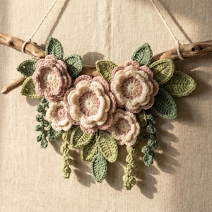 Könnte beinhalten: Handgefertigte Blumen-Wanddekoration mit gehäkelten Blüten in Rosa und Creme, akzentuiert mit grünen Blättern und hängenden Ranken. Die Blumen sind an einem natürlichen Ast befestigt, der an einer cremefarbenen Schnur hängt. Der Ast ist etwa 30 cm lang.