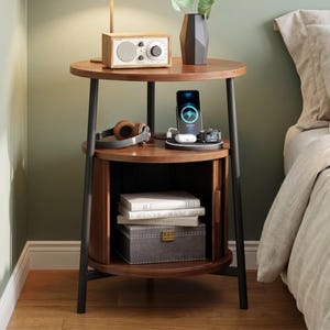 May include: A two-tiered, round wooden nightstand with black metal legs. The top shelf holds a small radio, a vase, and a lamp. The middle shelf has a charging station, headphones, and a smart watch. The bottom shelf has books and a storage box.