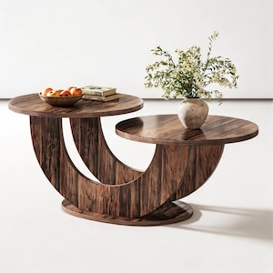 May include: A two-tiered coffee table with a dark brown wood grain finish and a curved, crescent-shaped base. The upper level holds a wooden bowl with several apples and two books, while the lower level supports a textured vase with white flowers.