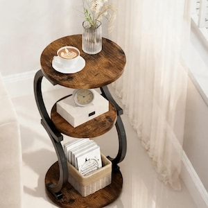 May include: A three-tiered side table with a dark brown, wood-grain finish. The table has a black metal frame and holds a cup of coffee, a white box, a small clock, and a basket of items. A vase of flowers sits on the top tier.