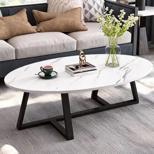 May include: Oval coffee table with a white marble-look top and black metal legs. A green teacup and saucer, a decorative deer figurine, and a vase of flowers are on the table. The table is in a living room setting.
