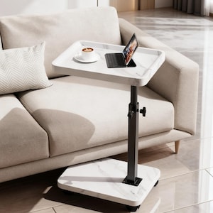 May include: A white marble-patterned laptop table with a black adjustable stand. A laptop and a cup of coffee sit on the table. The table is next to a beige sofa. The table has a rectangular top and a square base with wheels.