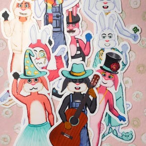 May include: A collection of whimsical rabbit character stickers, each dressed in unique outfits and accessories. The rabbits are depicted in various poses, some playing instruments, wearing hats, and holding props. The background is a light pink color.