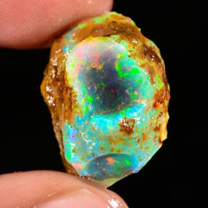 May include: A raw, unpolished opal gemstone featuring a translucent, iridescent center with vibrant flashes of neon green, electric blue, and soft pink. The stone is surrounded by a rough, textured, earthy brown matrix rock.