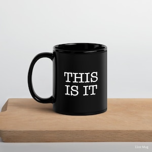 Puede incluir: Una taza de cerámica negra con el texto "THIS IS IT" en blanco, exhibida sobre una superficie de madera. La taza tiene un asa curva y una forma cilíndrica. La taza es de 325 ml.