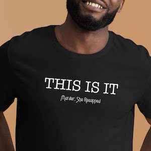 Puede incluir: Camiseta negra con el texto blanco "THIS IS IT" y el texto "Murder, She Reappeared" debajo. La camiseta está hecha de un material suave.