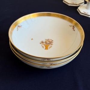 Może przedstawiać: Trzy białe porcelanowe miski ze złotymi obwódkami i wzorami koszy kwiatowych. Miski mają złoty brzeg z delikatnym wzorem. Wzór kosza kwiatowego jest w odcieniach brązu i złota. Miski są ułożone jedna na drugiej.
