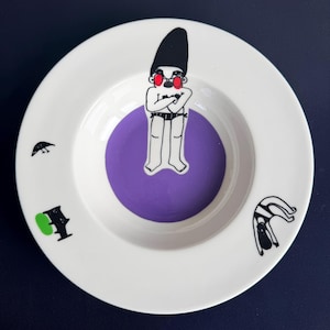 May include: White ceramic plate with a purple center and whimsical illustrations. The plate features a cartoon figure with a black hat and red sunglasses, along with other black and green graphic elements. A unique decorative piece.