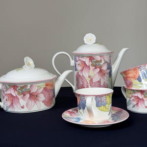 Peut inclure: Service à thé blanc avec motifs floraux, comprenant une théière, deux tasses, une soucoupe et un pot à crème. L'ensemble présente des motifs floraux roses, oranges et bleus. La théière et le pot à crème ont des couvercles blancs avec des boutons en forme de fleur.