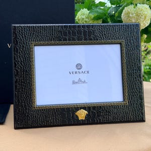 May include: Black rectangular picture frame with a textured finish and gold detailing. The frame features the Versace and Rosenthal logos. The frame is displayed on a beige surface.