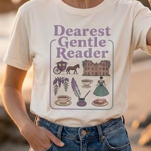 Puede incluir: Camiseta color crema con el texto "Dearest Gentle Reader" en morado, enmarcado por un borde decorativo. El gráfico incluye ilustraciones de un carruaje, un caballo, un edificio, un vestido y otros elementos. La modelo lleva vaqueros azules.
