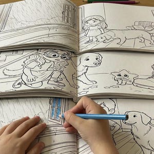 May include: A child's hand using a blue pencil to colour a page in a colouring book. The book shows illustrations of a girl, a dog, a star, and a boot. Coloured pencils and a pencil sharpener are on the wooden table.