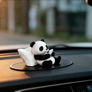 May include: A small panda figurine in black and white, depicted in a seated position while holding a white rectangular object. The panda rests against a white pillow, and the entire set is placed on a black oval-shaped textured mat.