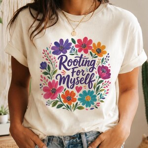 Puede incluir: Una camiseta de manga corta color crema con un diseño de corona floral circular en tonos rosa, morado, naranja y azul. El texto Rooting For Myself está impreso en el centro en una fuente cursiva morada junto a un pequeño corazón rojo.