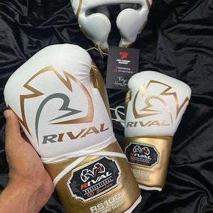 Könnte beinhalten: Weiß-goldenes Box-Set, bestehend aus Handschuhen und Kopfschutz. Die Handschuhe und der Kopfschutz zeigen das "RIVAL"-Logo und das Branding der "RS100"-Serie. Das Set ist für Sparring und den professionellen Einsatz konzipiert.