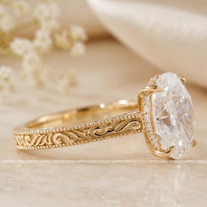 Op de afbeelding: Een gouden verlovingsring met een ovaal geslepen diamant. De ring heeft een gedetailleerde, gegraveerde band en een halo van kleinere diamanten rond de hoofdsteen. De ring is geplaatst tegen een zachte, neutrale achtergrond.