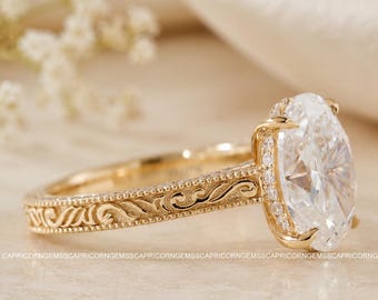 Peut inclure: Une bague de fiançailles en or avec un diamant de taille ovale. La bague présente une bande gravée détaillée et un halo de petits diamants autour de la pierre principale. La bague est posée sur un fond doux et neutre.