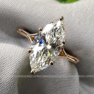 May include: A marquise-cut diamond ring with a rose gold band. The large, clear diamond is the focal point, set in a delicate, pronged setting. The ring is displayed on a soft, grey fabric, with a blurred green background.