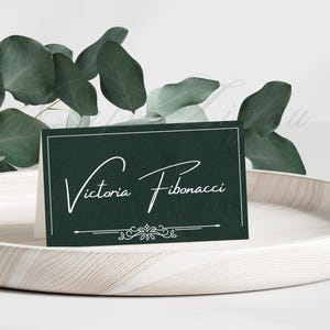 May include: A dark green place card with the name "Victoria Fibonacci" in elegant white script. The card has a white border and decorative elements at the bottom. Eucalyptus leaves and a wooden tray provide a natural backdrop.