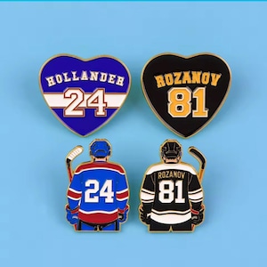 May include: Two heart-shaped enamel pins featuring the names Hollander 24 and Rozanov 81, alongside two pins depicting hockey players from the back wearing jerseys with the same names and numbers, including hockey sticks and helmets.