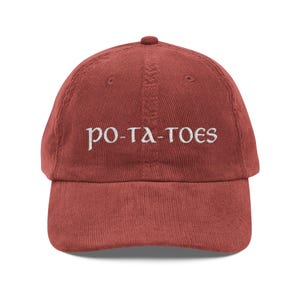 May include: A brick-red corduroy baseball cap with the word "po-ta-toes" embroidered in white. The cap has a curved brim and an adjustable strap at the back. A casual, everyday accessory.