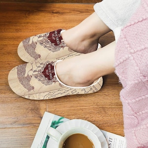 May include: A pair of beige slippers featuring a map design with the text "The Marauders Map" in burgundy. The slippers are on a wooden floor, with a cup of coffee and a magazine in the foreground.