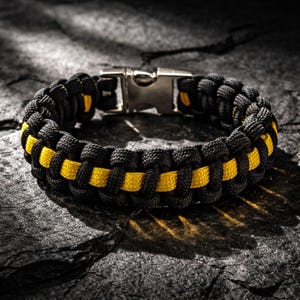 May include: Black and yellow paracord bracelet with a stainless steel clasp. The bracelet is made of tightly woven black cord with a yellow stripe running through the center. The clasp is silver and securely fastens the bracelet.