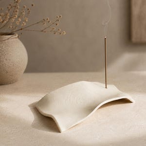 Puede incluir: Un porta-incienso de cerámica color crema, curvado con una superficie lisa y ondulada. Una varilla de incienso fina y marrón está insertada en el soporte, emitiendo un tenue hilo de humo que se eleva verticalmente sobre el objeto.