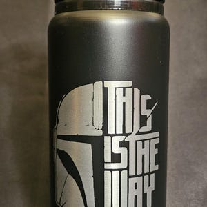 May include: Black water bottle with a flip-top lid. The bottle features a silver graphic of a Mandalorian helmet and the phrase "THIS IS THE WAY" in a stylized font. The bottle is cylindrical.