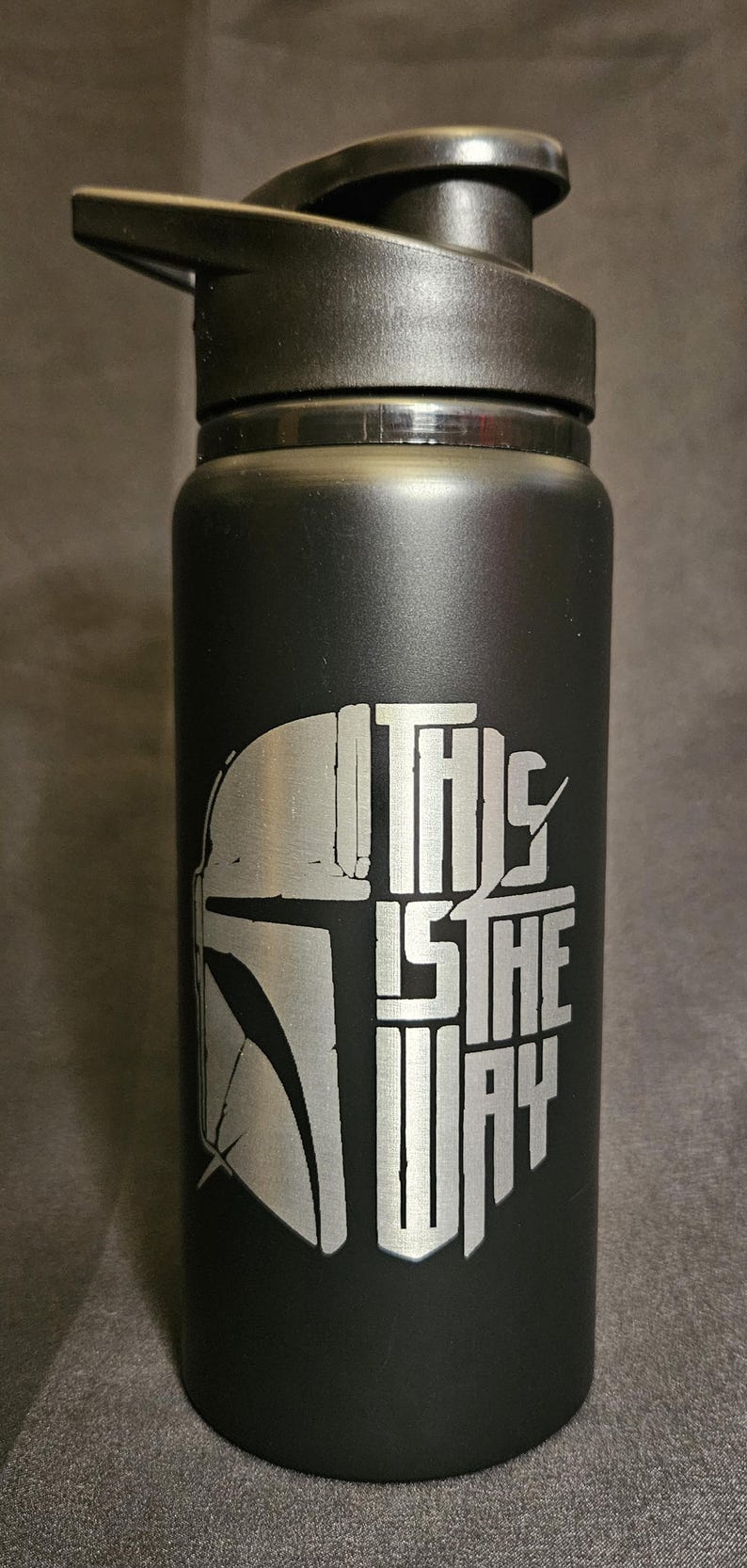 May include: Black water bottle with a flip-top lid. The bottle features a silver graphic of a Mandalorian helmet and the phrase "THIS IS THE WAY" in a stylized font. The bottle is cylindrical.
