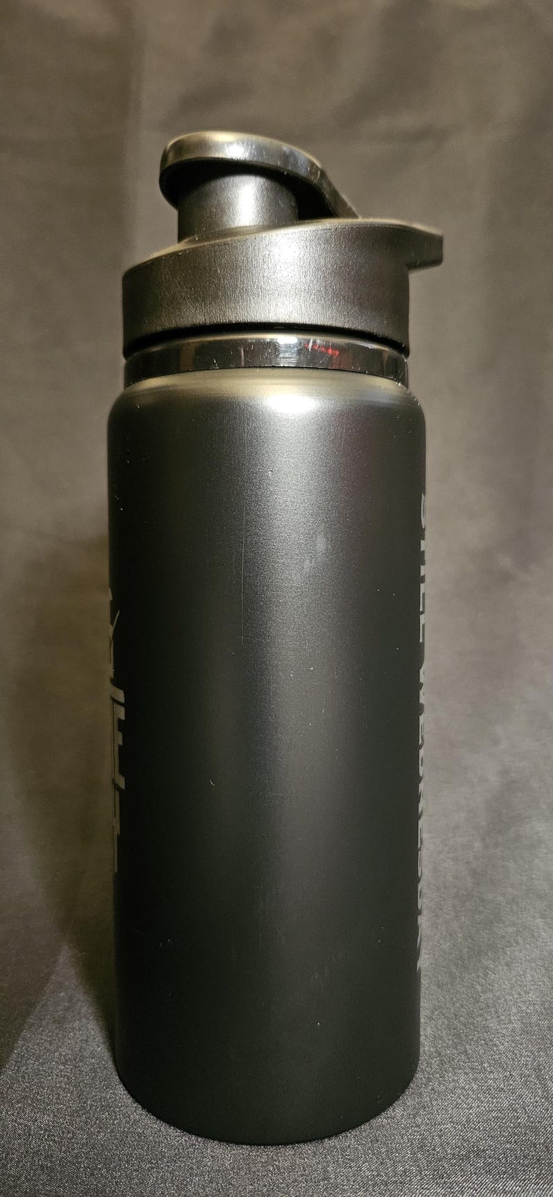 May include: Black insulated water bottle with a flip-top lid. The bottle has a matte finish and a cylindrical shape. Text is visible on the side. Ideal for sports, travel, or everyday hydration.
